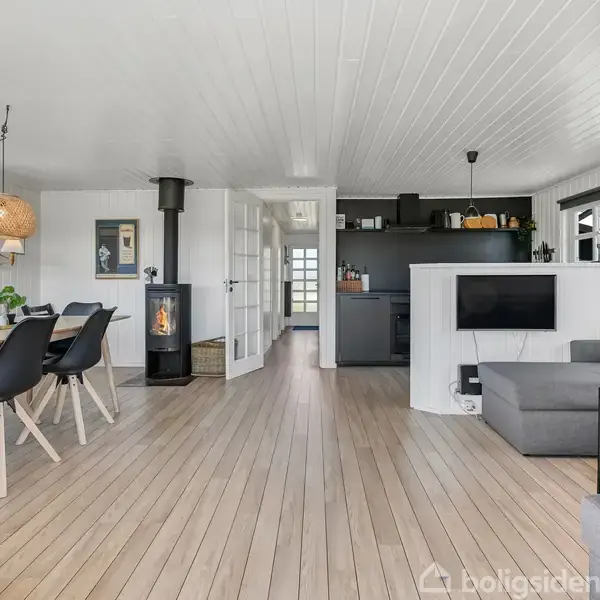 En moderne stue med lyse trægulve. En sofa med puder står foran et tv på væggen. Et spisebord med stole og en brændeovn er i baggrunden.