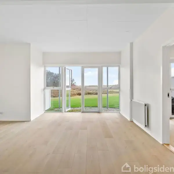Et lyst, tomt rum med trægulv leder ud mod en stor terrasse via glasdøre. Gennem dørene ses en græsplæne og et åbent landskab. En dør til højre fører til et andet rum.