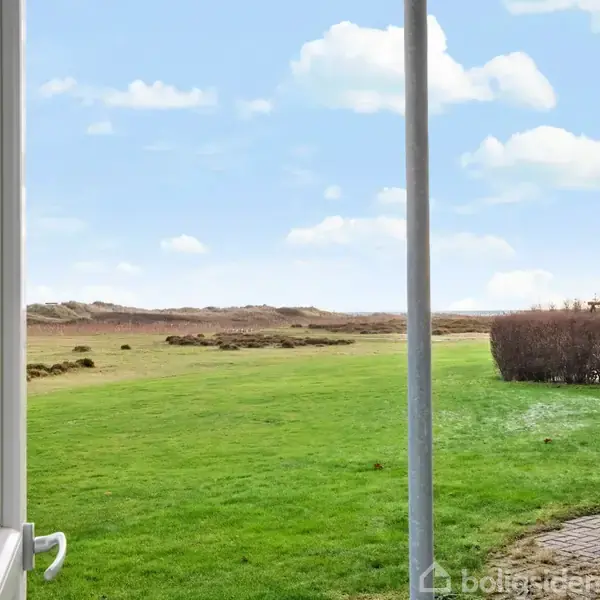 En åben glasdør viser en grøn græsplæne, der strækker sig mod fjerne klitter under en blå himmel med lette skyer. En brolagt sti ses til højre.