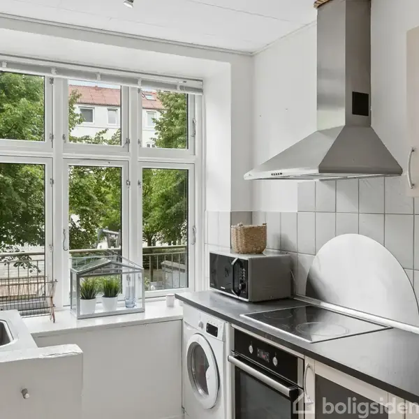 Håndvask med ternet flisebaggrund og spejl ovenover. Grå håndklæde hænger på væggen. Toilettet ses delvist i højre side. Rør løber langs væggen i baderumsmiljøet.