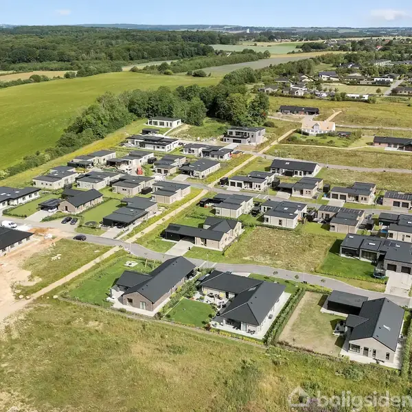 Boligområde med moderne huse spredt i et gittermønster. Græsplæner omgiver husene, og åbne marker strækker sig i baggrunden. Skovstrækninger og en klar himmel omkranser området.