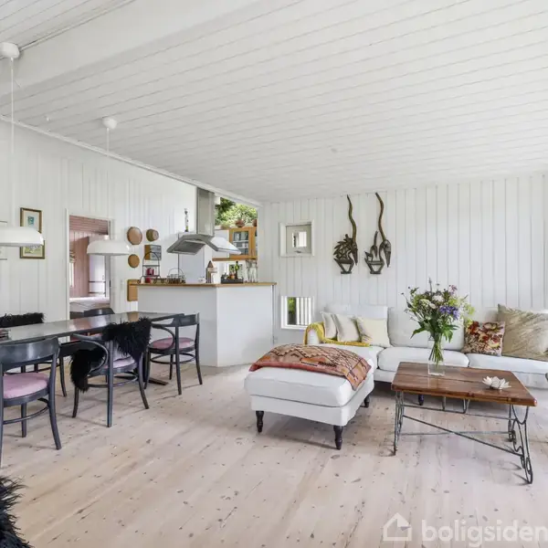 Stue med spisebord og stole til venstre, en hvid sofa og sofabord i midten, vægpynt og planter. Rummet har lyse trævægge og vinduer med naturligt lys.