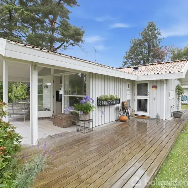 Sommerhus med en træterrasse omkranset af grøn vegetation, blomster og havekrukker. Beliggende i en frodig have med høje træer og blå himmel i baggrunden.