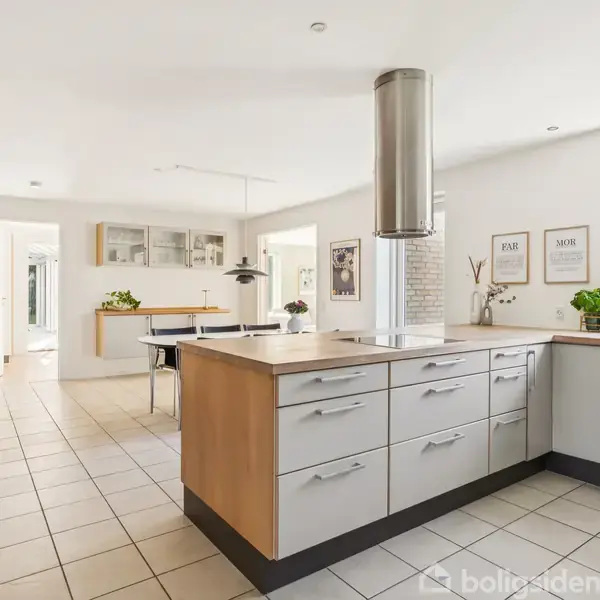 Et moderne køkken med en stor køkkenø, hvide skabslåger, stålventilation over kogepladerne og et åbent spiseområde med et bord i baggrunden. Lyse fliser dækker gulvet.