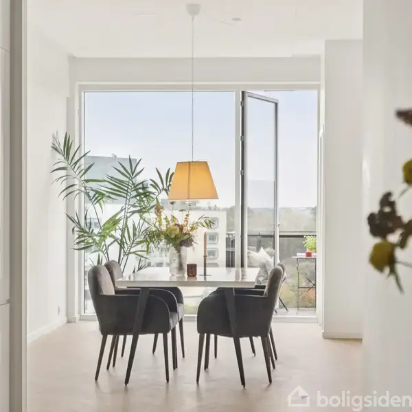 Fire polstrede stole står rundt om et spisebord med en hængende lampe ovenover. Store glasdøre åbner ud mod en altan. Grønne planter står ved siden af bordet.