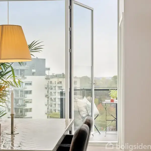 Et stilfuldt spisebord med blomsterdekoration står i et lyst rum. En åben dør fører ud til en balkon med udsigt til moderne bygninger og en klar himmel.