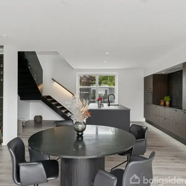 En moderne spisestue indeholder et rundt, sort bord med fire stole. Bagved er et åbent køkken med en stor bordplade og en trappe til venstre ved en vinkælder.