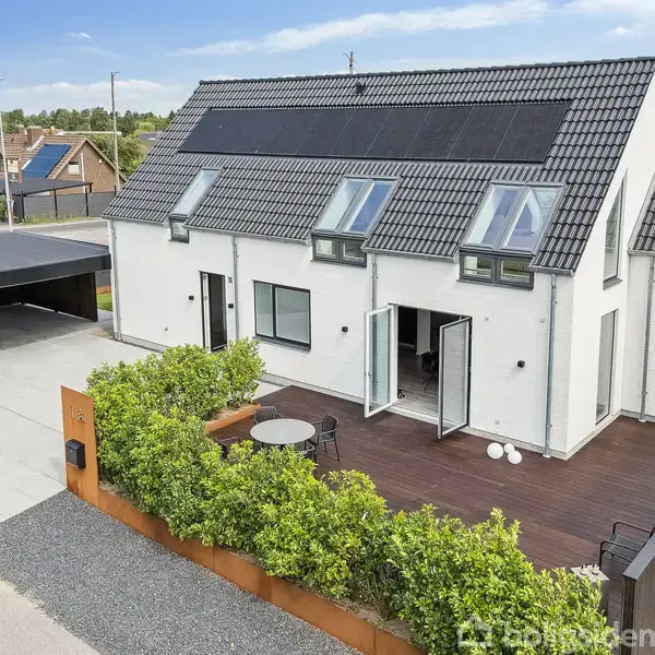 Hus med solpaneler på taget, åbne terrassedøre, beplantet have og tilhørende carport. Beliggende i et roligt boligområde med flere lignende huse i nærheden.