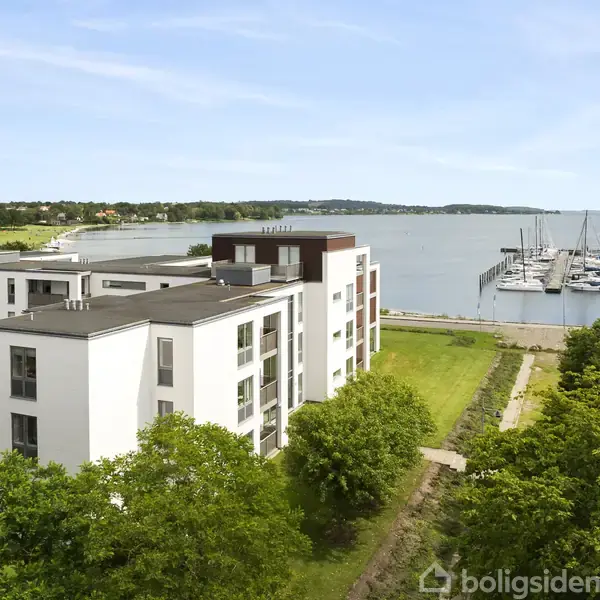 Bygningen står stille med et fladt tag og hvide facader, omgivet af grønne træer ved vandet. I baggrunden ses en marina med både fortøjet ved molen.