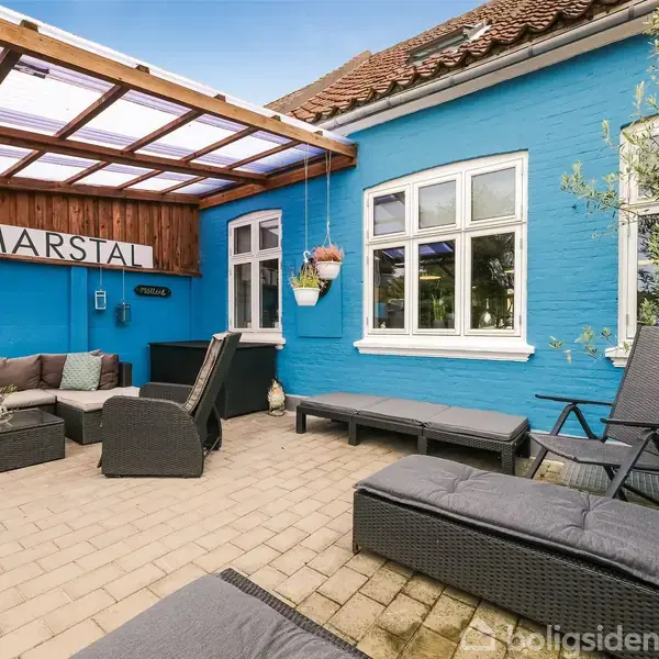 Køkken med grønne skabe, sort emhætte og ovn, glaslågeskab med porcelæn. Lyst rum med trægulv, hvide vægge og store vinduer. En dør fører til et andet rum.