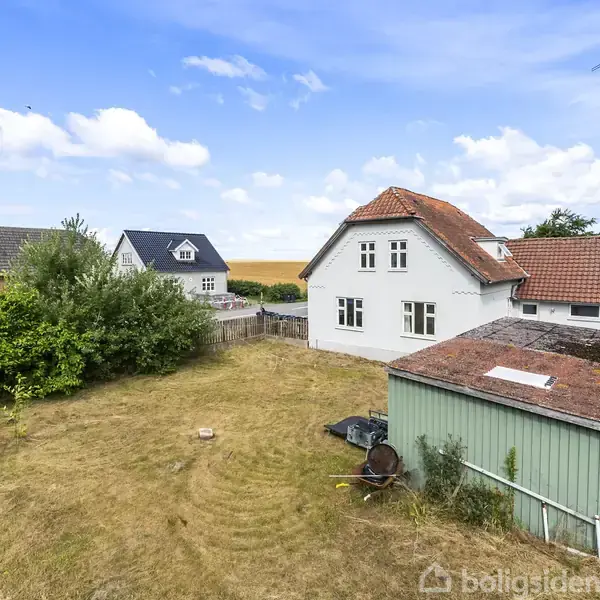 Hus med rødt tag står i en stor, åben have omgivet af buske og træer. To andre huse ses i baggrunden under en blå himmel med skyer.