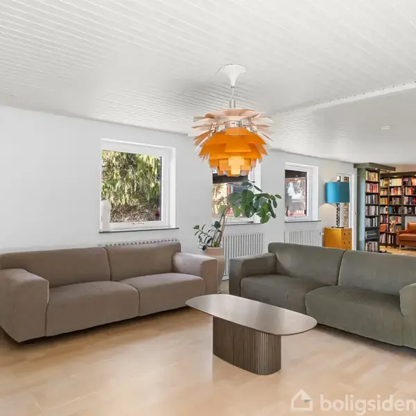 To sofaer i en lys stue, står over for et lavt sofabord, omgivet af vinduer og en stor bogreol. En orange loftslampe hænger ned fra loftet.