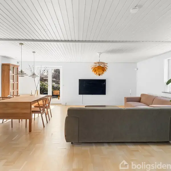 Stue med sofa og stort TV på væggen. Spisebord med stole står til venstre, mens to hængelamper hænger over. Store vinduer giver masser af lys. Maleri og plante dekorerer rummet.