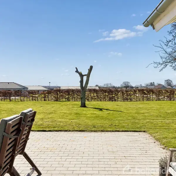 En terrasse med træmøbler står ved siden af et åbent græsareal med en skulpturelt beskåret træstamme. I baggrunden ses en hæk og bygninger under en klar blå himmel.