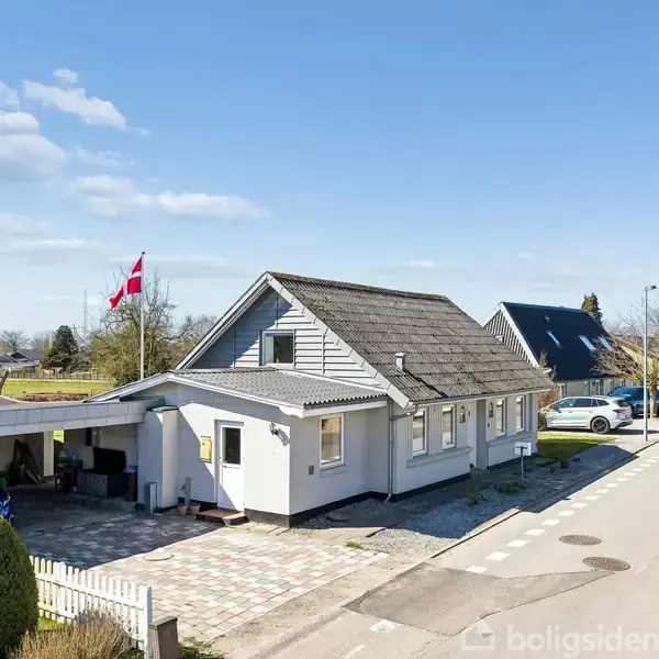 Hus med grå tag og hvid facade står ved vejen, omgivet af haver. I forgrunden ses en carport og dansk flag, mens vejen har parkerede biler og grønne områder.