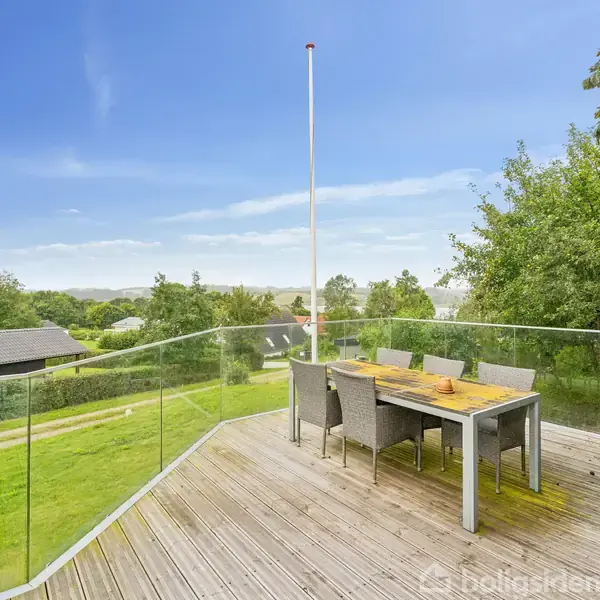 Terrassebord med seks stole står på en træterrasse. Det er omkranset af glasrækværk, placeret i en grøn have med udsigt over et roligt, landligt område og huse i baggrunden.