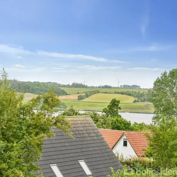 Et hustag ligger stille omgivet af grønne træer, med en udsigt over en dal med marker og en flod i baggrunden under en klar blå himmel.