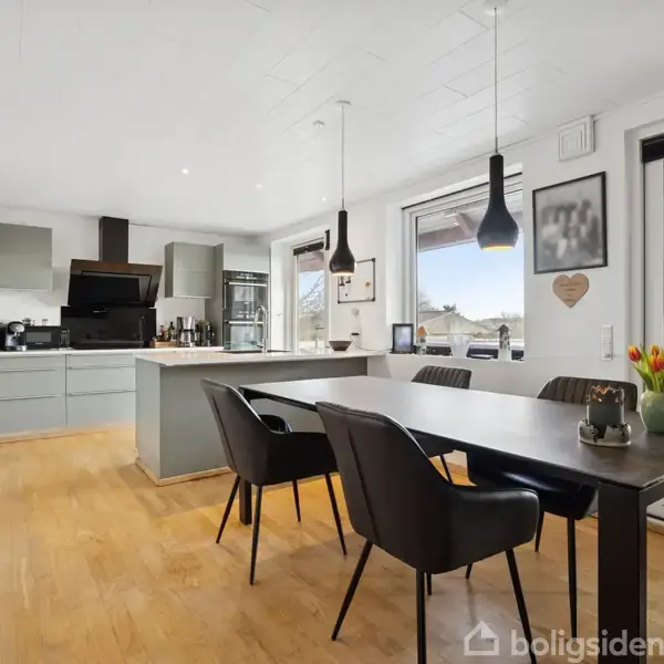 Et moderne køkken med træbord, sorte stole, køkkenø og grå skabe. Bordet har en vase med tulipaner. Store vinduer og glasdøre giver udsigt og lys.