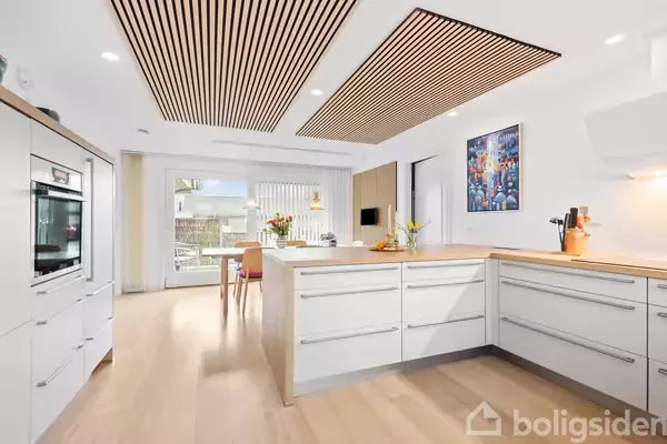 Et lyst køkken med moderne, hvide skabe og træbordplader. En frugtskål og blomsterdekoration står på øen, og et farverigt maleri hænger på væggen. I baggrunden ses en spiseplads ved et stort vindue.