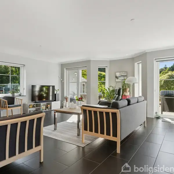 En lys stue med moderne møbler, hvor to sofaer og en lænestol er omkring et sofabord. Et fjernsyn står på et lavt skab. Store vinduer slipper sollys ind.