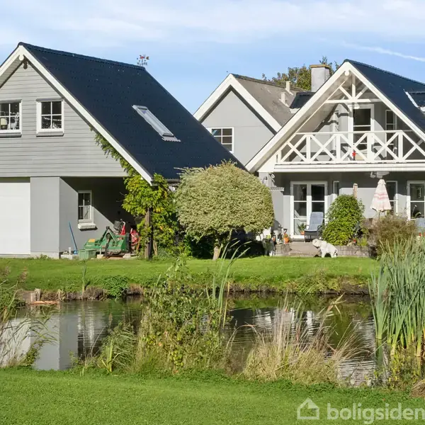 Et gråt hus med sort tag står i sollys. Det har en stor garage og balkon. Foran huset er en dam omgivet af grønt græs og træer.