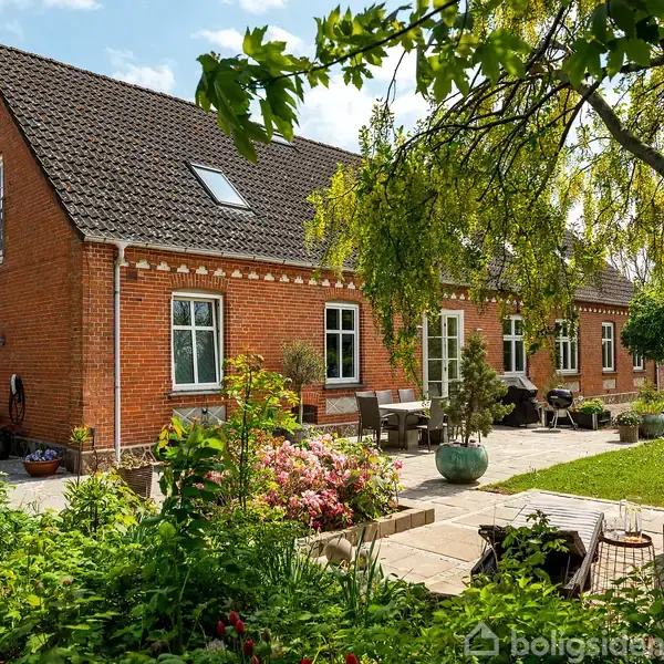 Murstenshus med hvide vinduer står stille i en frodig have med havemøbler og blomsterbede. Sollys skinner gennem træernes grene og skaber en fredfyldt atmosfære.