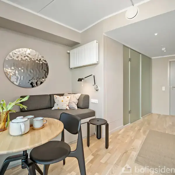 Et lille stuebord med kopper og en blomst står foran en grå sofa. Rummet har trægulv, en rund vægdekoration og døren til en gang i baggrunden.