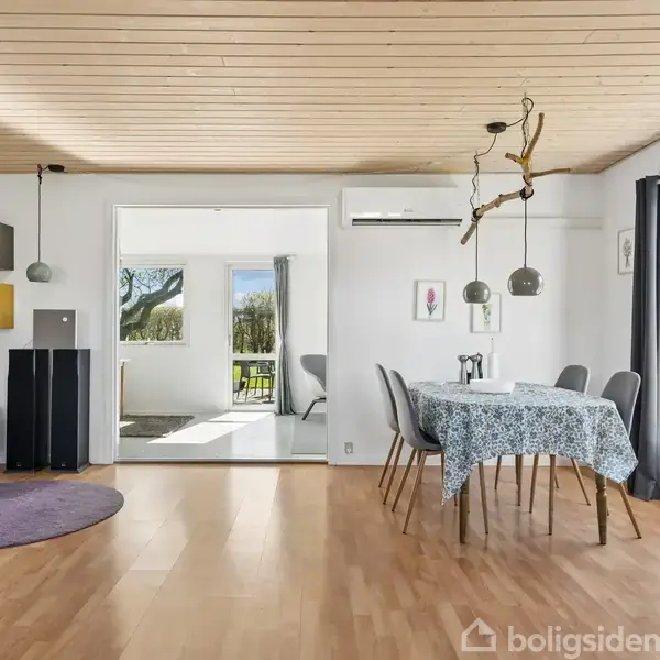 Stue med spisebord dækket med dug, flankeret af stole. Træloft og parketgulv. Sofa til venstre, vindue med udsigt til have. Lampe og klimaanlæg hænger over bordet. Ingen tekst.