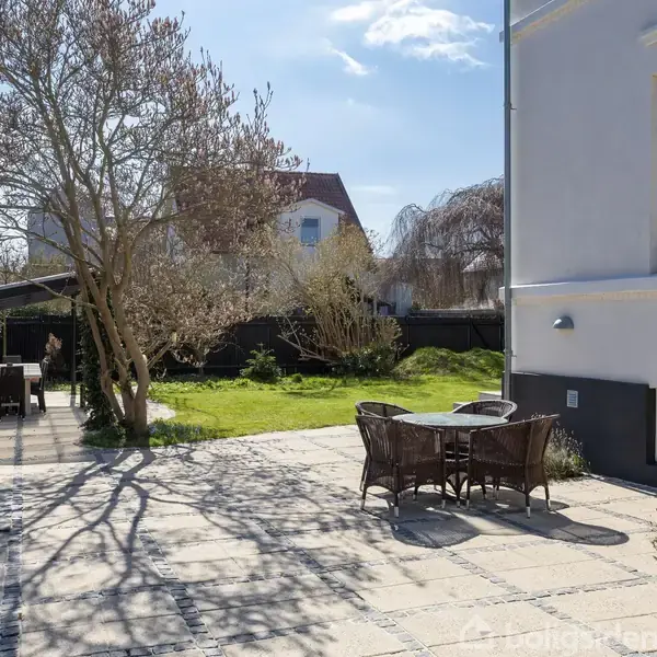 Et rundt havebord med stole står på en flisebelagt terrasse. I baggrunden ses en græsplæne, træer og en bygning med hvide vægge under en klar blå himmel.