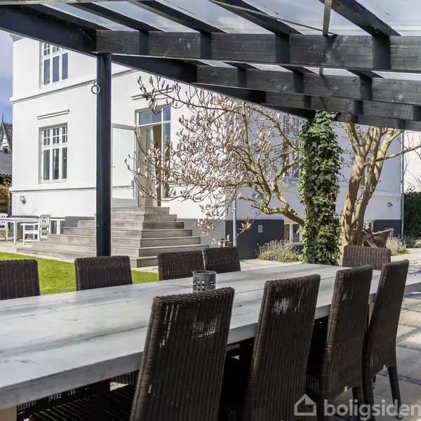 Langt spisebord med mørke kurvestole stående på en terrasse under et glasoverdækning. I baggrunden er der en hvid to-etagers bygning, have og træer.
