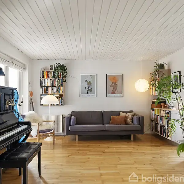 Klaver ved siden af sort taburet i en stue. Sofa med puder står op ad væggen. Reoler med bøger flankerer rummet. Lys træplankegulv og loft i lyst træ. Planter dekorerer hjørnerne.