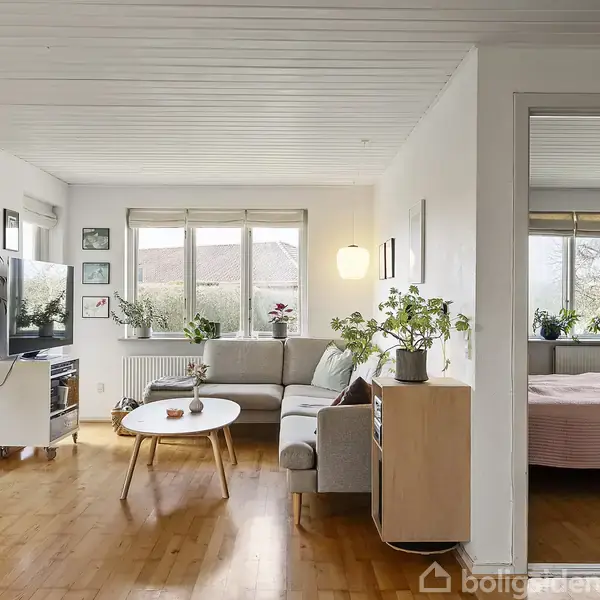 Stue med grå sofa og hvidt rundt sofabord foran stort vindue. Planter og billeder dekorerer rummet. En åben dør fører til et soveværelse med seng.