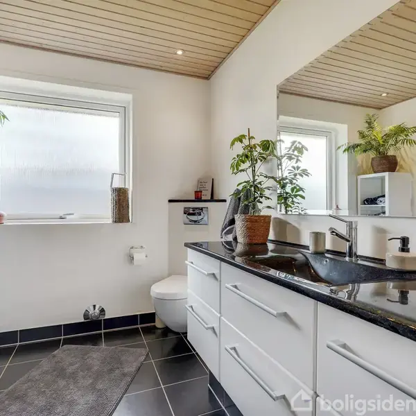 Badeværelse med hvidt, moderne møblement, sort bordplade og spejl. Planter pynter rummet. Der er et vindue ved toilettet, sorte fliser på gulvet og et hvidt træloft.