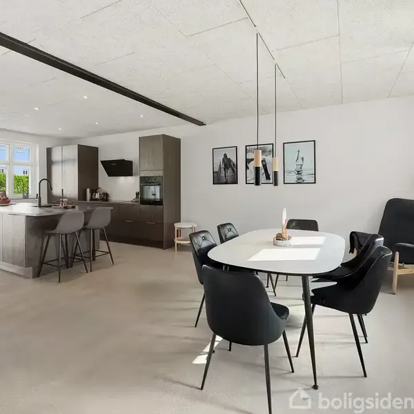 Et moderne køkkenalrum med åbent køkken og spisebord. En køkkenø med barstole står til venstre; to billeder hænger på væggen bagved spisebordet. En stor stol i hjørnet. Ingen tekst fundet.
