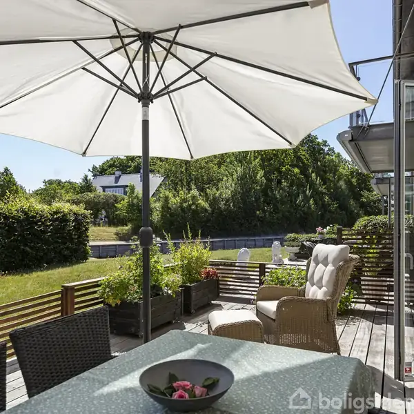 En stor hvid parasol står åben over en terrasse med spisebord og stole. Terrassen er omgivet af grønne planter og en velplejet have i solrigt vejr.