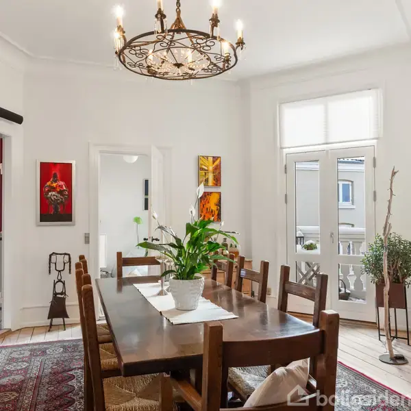 Et elegant spisebord i mørkt træ med stole omkring står i midten af et lyst rum. En stor lysekrone hænger over bordet. Kunstværker pryder væggene.