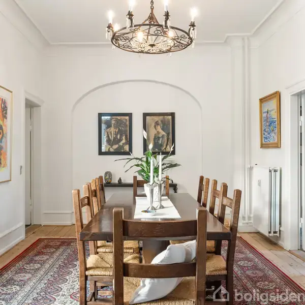 Spisebord med seks stole står på et orientalsk tæppe i en lys spisestue. Over bordet hænger en lysestage, væggene har kunstværker og en vitrine viser glas.