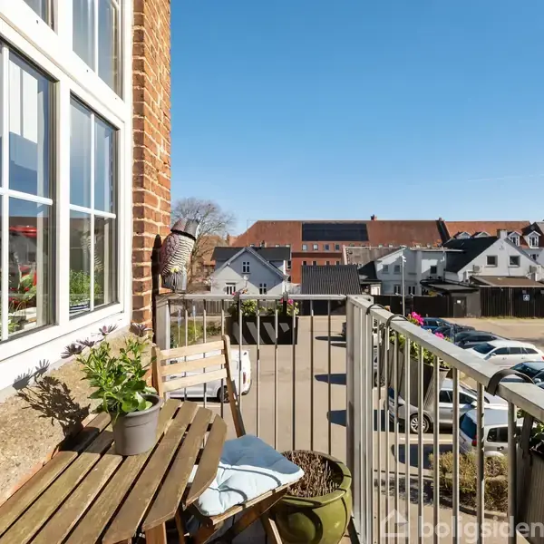 En lille altan med en træbænk og blomsterkrukker står ved siden af et murstenshus. I baggrunden ses en parkeringsplads og rødt tegltagede bygninger under en klar blå himmel.