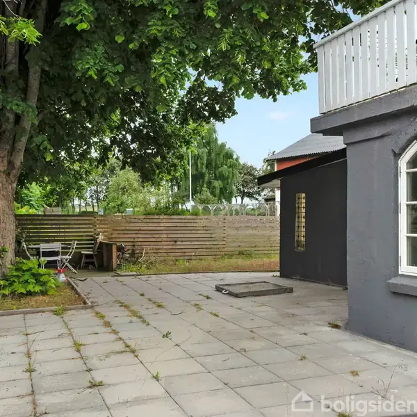 Terrasse omgivet af træer og et hegn. Et gråt hus med buede vinduer står til højre. På terrassen er der et bord og stole under et stort træ.