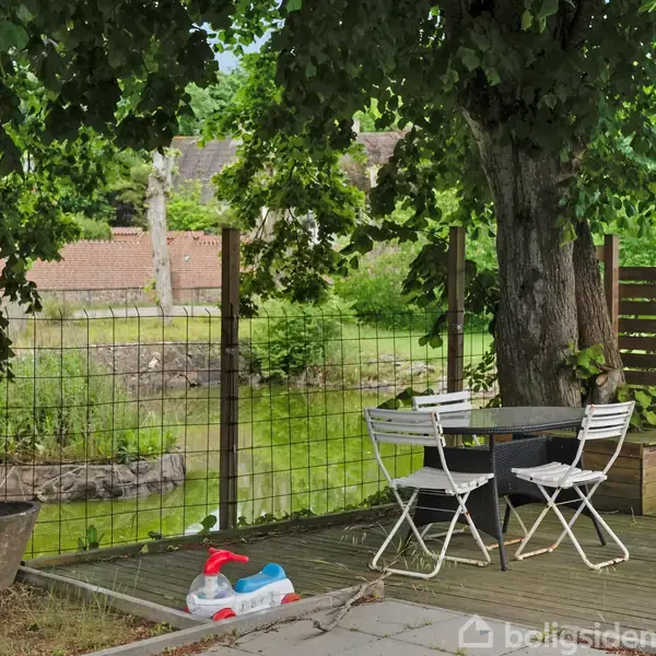 Et sæt hvidmalede havemøbler står på en træterrasse under et stort træ. En legetøjsbil er placeret ved siden af, og et trådhegn omgiver området med grønne planter i baggrunden.