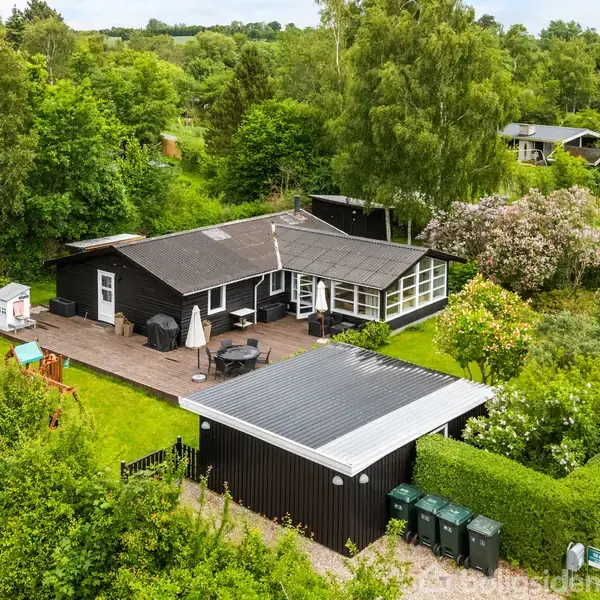 Hus med mørkt tag, omgivet af grønne træer og buske, står ved siden af en træterrasse med havemøbler og parasol. Til højre er der en grusvej og skraldespande.