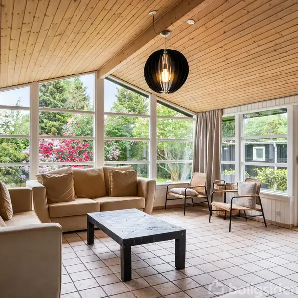 Stue med beige sofa og bord, stående på flisegulv. Store vinduer med udsigt til frodigt grønt haveområde. Loft af træ og sort pendellampe giver hyggelig atmosfære. Ingen tekst.
