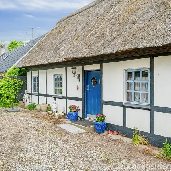 Et bindingsværkshus med stråtag har en blå dør og små vinduer. Foran står blå krukker med blomster. Der er statuer og en udendørslampe ved indgangen.