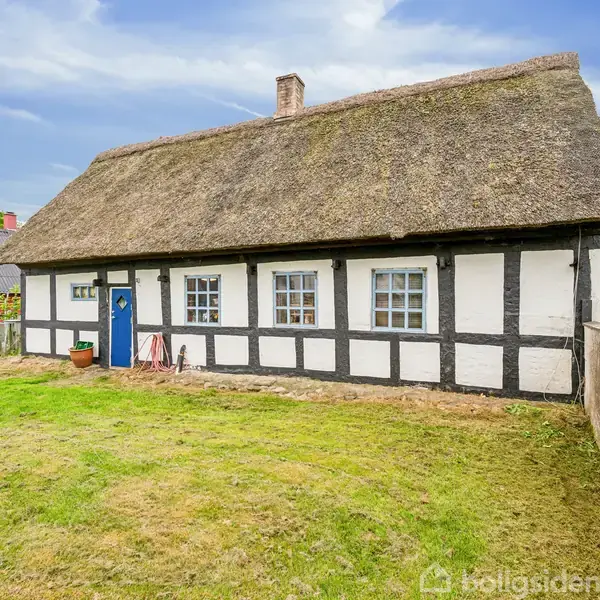 Bindingsværkshus står ubenyttet med stråtag, omgivet af en velplejet græsplæne i et landsbyområde med flere charmerende, ældre bygninger.
