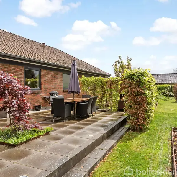Hus med terrasse, hvor et bord og stole står under en parasol. Terrassen er omgivet af buske og blomster, og der er en græsplæne i baggrunden under en blå himmel.