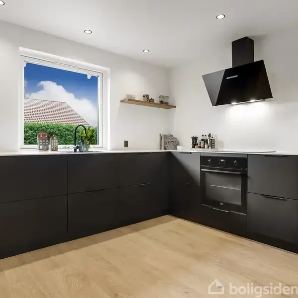 Et moderne køkken med sorte skabe, indbygget ovn og emhætte. Der står en kaffemaskine på bordet ved et vindue med udsigt til en murstensbygning og træer udenfor.