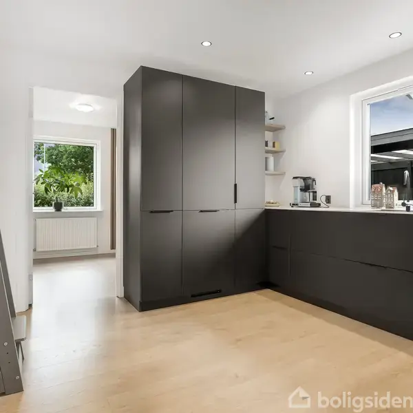 Et moderne køkken med sorte skabe er stille i et lyst rum. Der er en stige til venstre, og vinduer viser et grønt, udendørs område og en velplejet have.