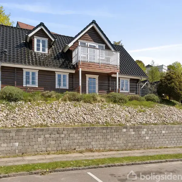 Hus med sort tag og hvid altan står på en hævet grund med sten og buske. Omkringliggende huse og grønne træer er synlige.