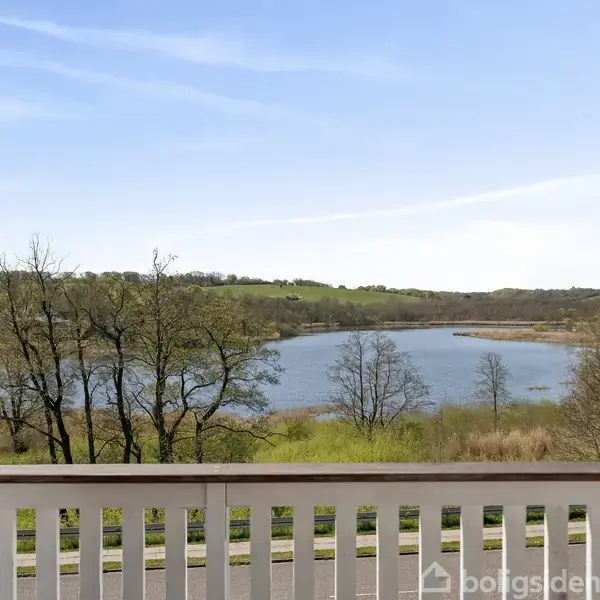 En åben altandør med udsigt over en sø omgivet af grønne bakker og træer. Klar blå himmel og en rolig atmosfære.