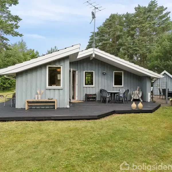 Gråt træhus med sort terrasse, omgivet af græsplæne og skov. Terrassen har havemøbler, og huset har en antenne på taget.